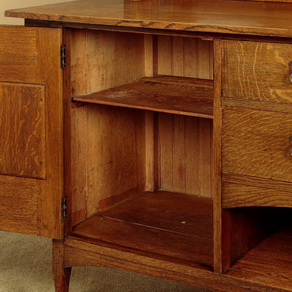 An Arts and Crafts quarter-sawn oak dresser featuring a mirrored top structure with small display shelves, inlaid floral details, hammered copper strap hinges, two drawers, and a lower cupboard. View X