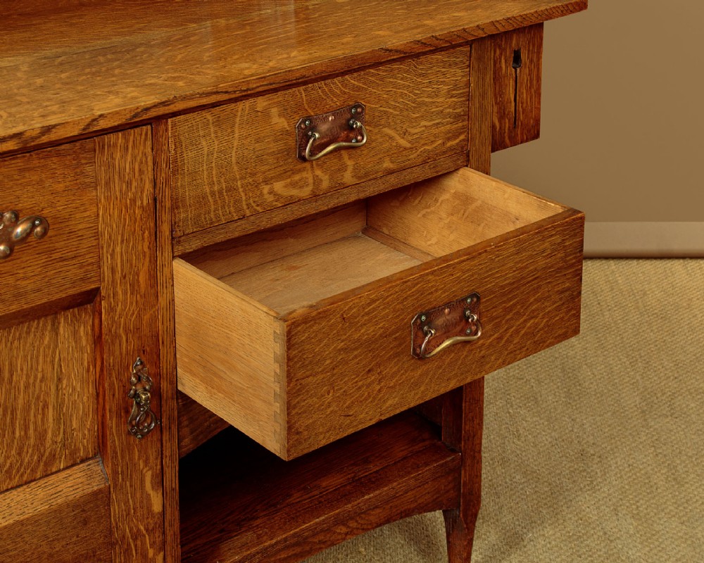 An Arts and Crafts quarter-sawn oak dresser featuring a mirrored top structure with small display shelves, inlaid floral details, hammered copper strap hinges, two drawers, and a lower cupboard. View XII