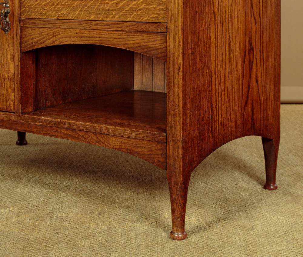 An Arts and Crafts quarter-sawn oak dresser featuring a mirrored top structure with small display shelves, inlaid floral details, hammered copper strap hinges, two drawers, and a lower cupboard. View XIII