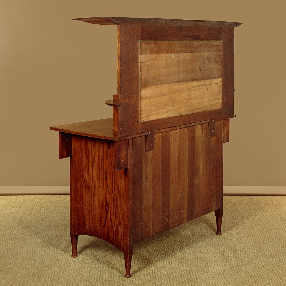 An Arts and Crafts quarter-sawn oak dresser featuring a mirrored top structure with small display shelves, inlaid floral details, hammered copper strap hinges, two drawers, and a lower cupboard. View XV