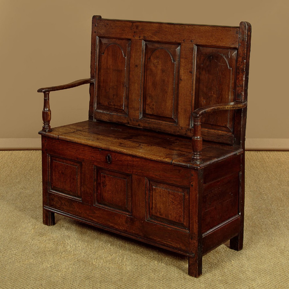 A dark oak box settle featuring a three-panel back with subtle arched top mouldings, curved armrests on turned supports, and a hinged seat opening to storage.
