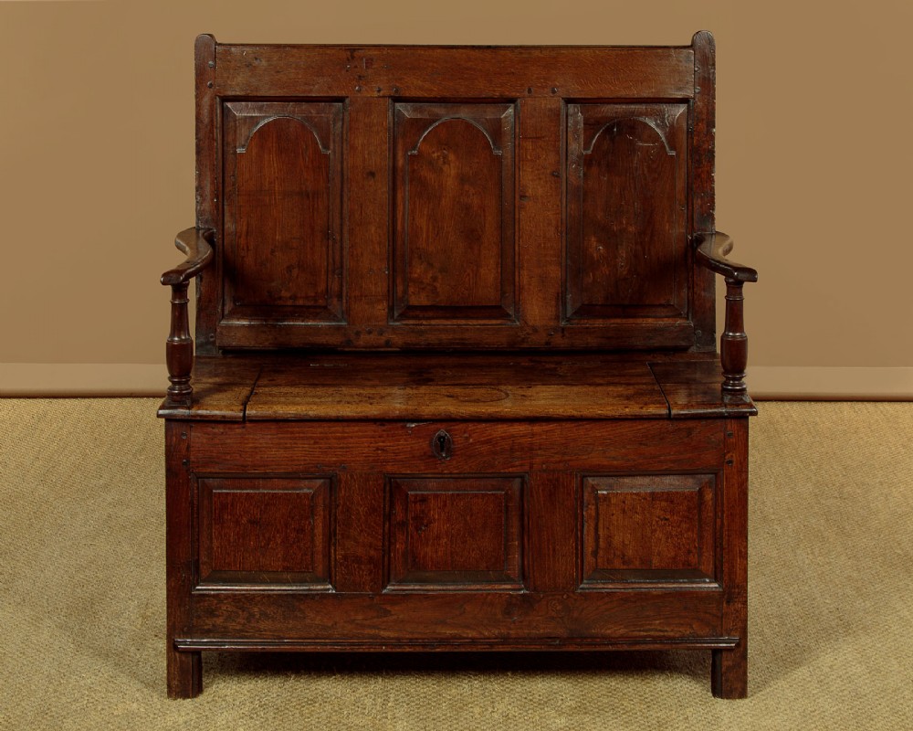 A dark oak box settle featuring a three-panel back with subtle arched top mouldings, curved armrests on turned supports, and a hinged seat opening to storage. View II
