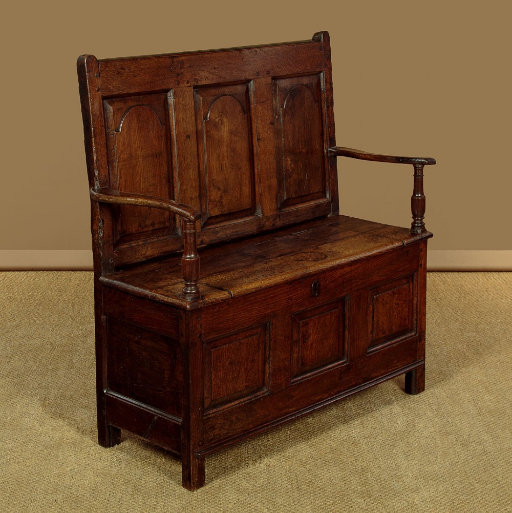 A dark oak box settle featuring a three-panel back with subtle arched top mouldings, curved armrests on turned supports, and a hinged seat opening to storage. View III