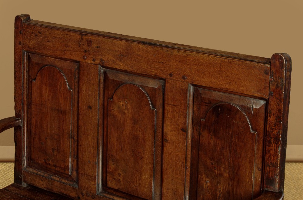 A dark oak box settle featuring a three-panel back with subtle arched top mouldings, curved armrests on turned supports, and a hinged seat opening to storage. View IV