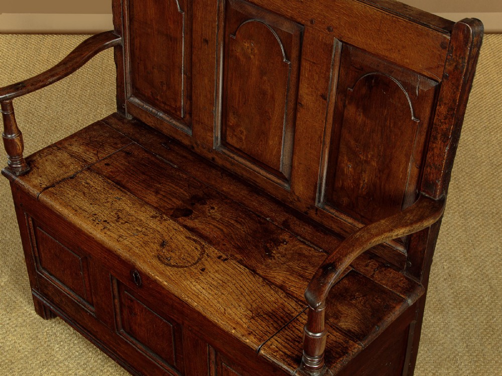A dark oak box settle featuring a three-panel back with subtle arched top mouldings, curved armrests on turned supports, and a hinged seat opening to storage. View VI