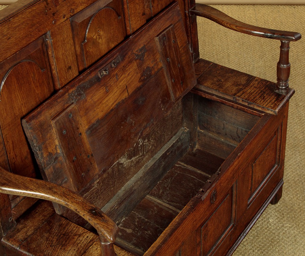 A dark oak box settle featuring a three-panel back with subtle arched top mouldings, curved armrests on turned supports, and a hinged seat opening to storage. View VII