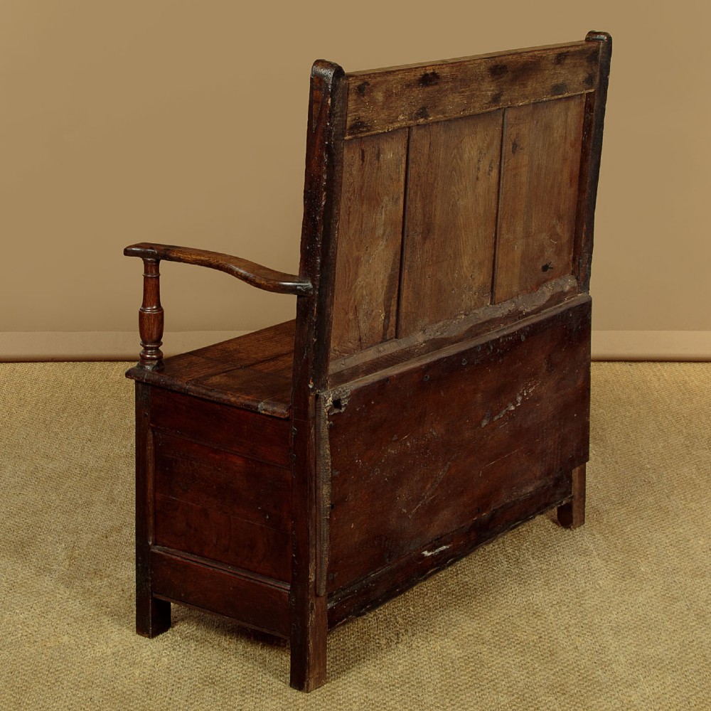 A dark oak box settle featuring a three-panel back with subtle arched top mouldings, curved armrests on turned supports, and a hinged seat opening to storage. View XII