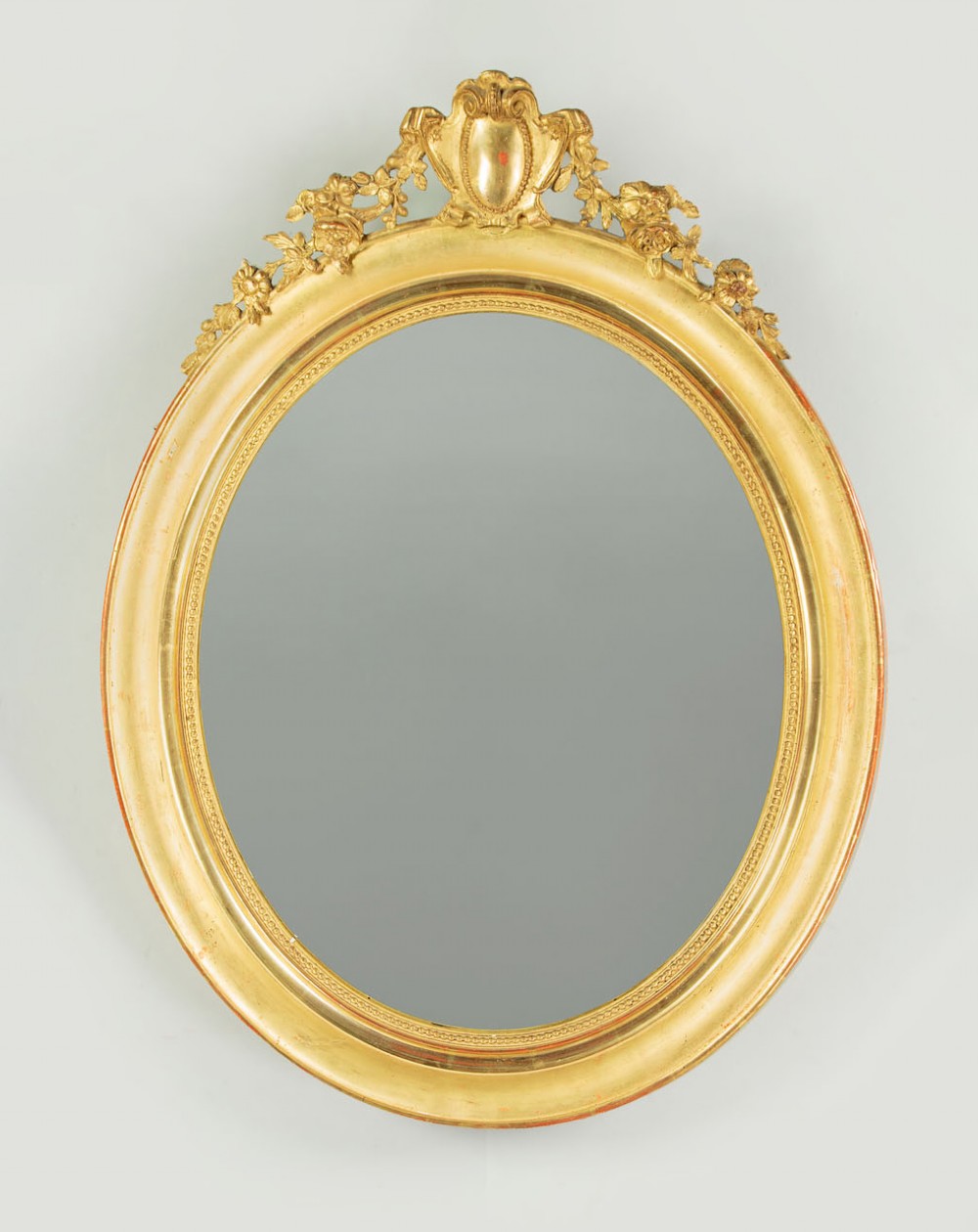 An antique oval giltwood wall mirror featuring a moulded frame with a beaded inner border and an ornamental cresting with floral swags.