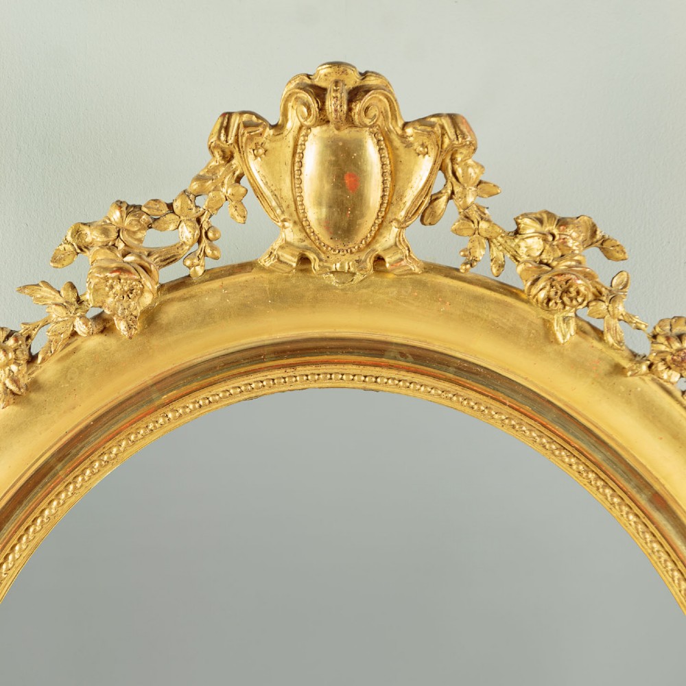 An antique oval giltwood wall mirror featuring a moulded frame with a beaded inner border and an ornamental cresting with floral swags. View III