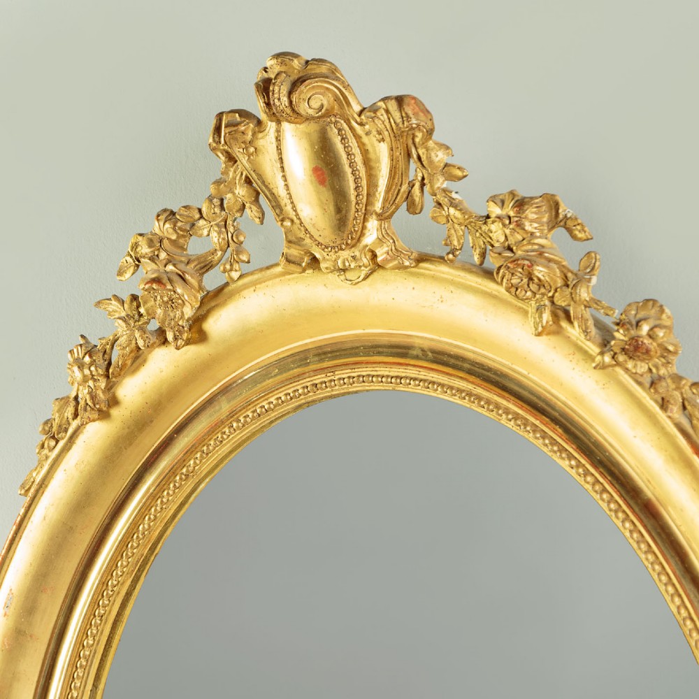 An antique oval giltwood wall mirror featuring a moulded frame with a beaded inner border and an ornamental cresting with floral swags. View IV