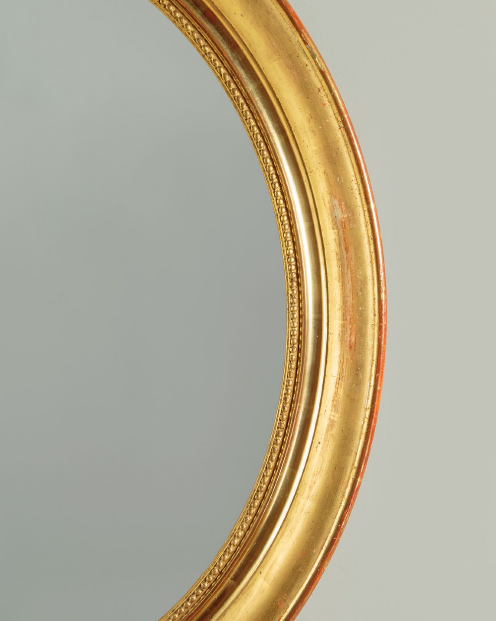 An antique oval giltwood wall mirror featuring a moulded frame with a beaded inner border and an ornamental cresting with floral swags. View V