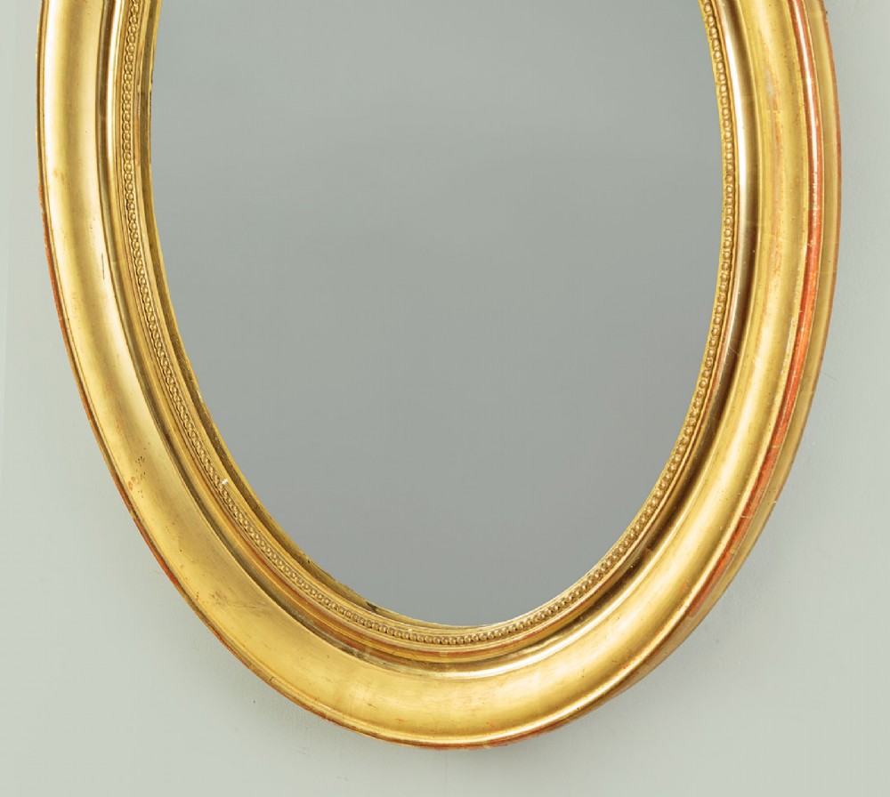 An antique oval giltwood wall mirror featuring a moulded frame with a beaded inner border and an ornamental cresting with floral swags. View VI