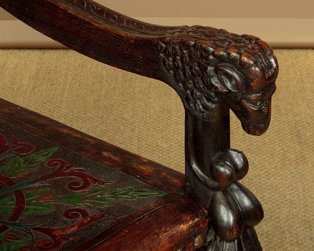 A tall, dark carved oak Renaissance Revival throne chair featuring a high back with a painted heraldic leather panel, lion head terminal armrests, and brass stud trim. View IX