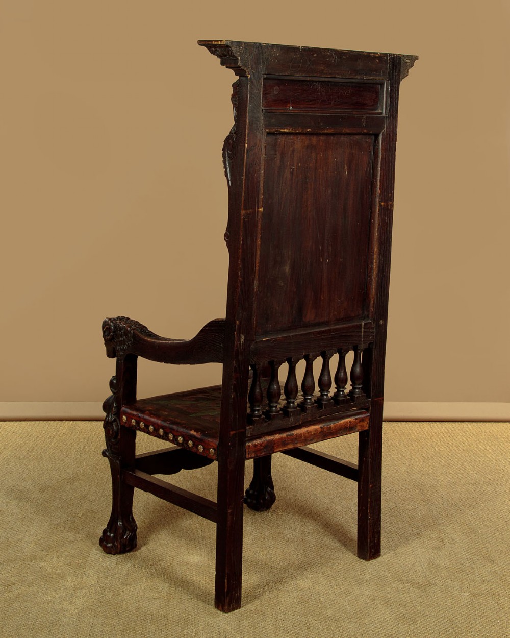 A tall, dark carved oak Renaissance Revival throne chair featuring a high back with a painted heraldic leather panel, lion head terminal armrests, and brass stud trim. View XII