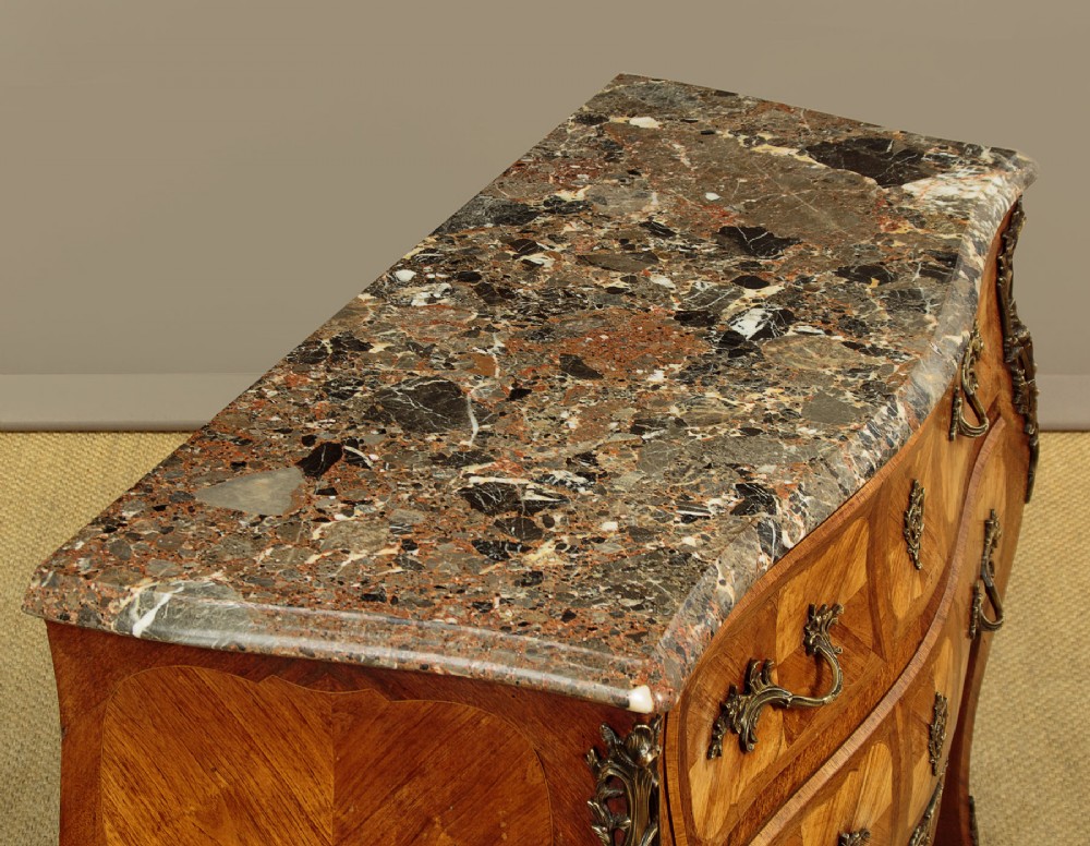 A 1930s Louis XV style bombe commode featuring a variegated grey marble top, parquetry wood veneer drawers, and ornate brass mounts. View V