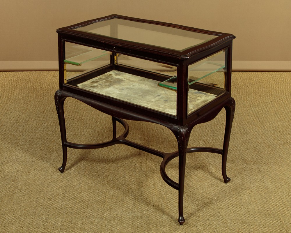 A dark mahogany glass-enclosed bijouterie display table featuring cabriole legs, curved stretchers, glazed side panels, and internal glass shelves.