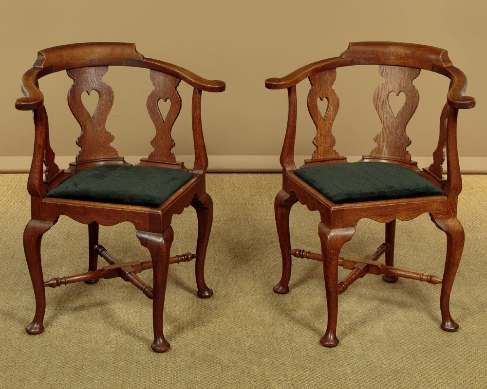 A pair of dark oak corner armchairs featuring pierced heart splats, cabriole legs, turned stretchers, and black upholstered seat cushions.