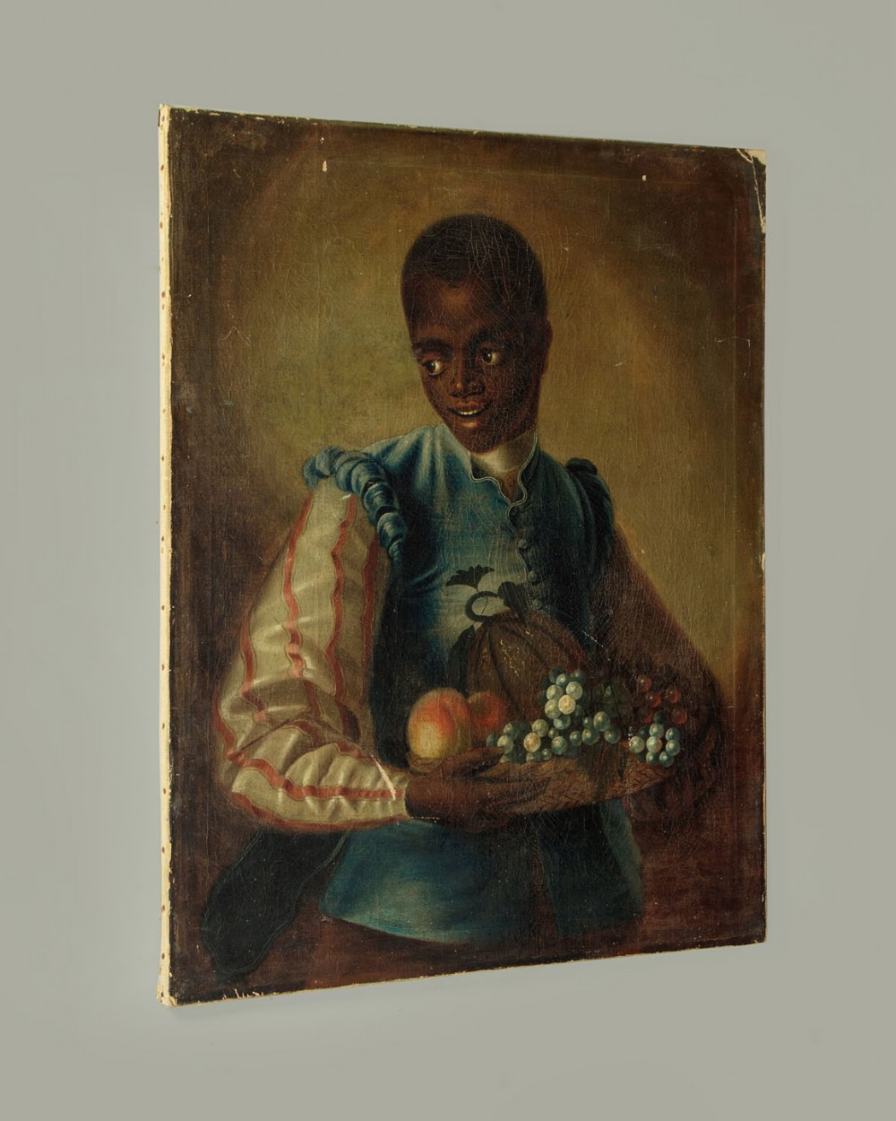 A Georgian style oil painting depicting a young servant holding a fruit basket containing grapes and a melon, dressed in a blue waistcoat. View II