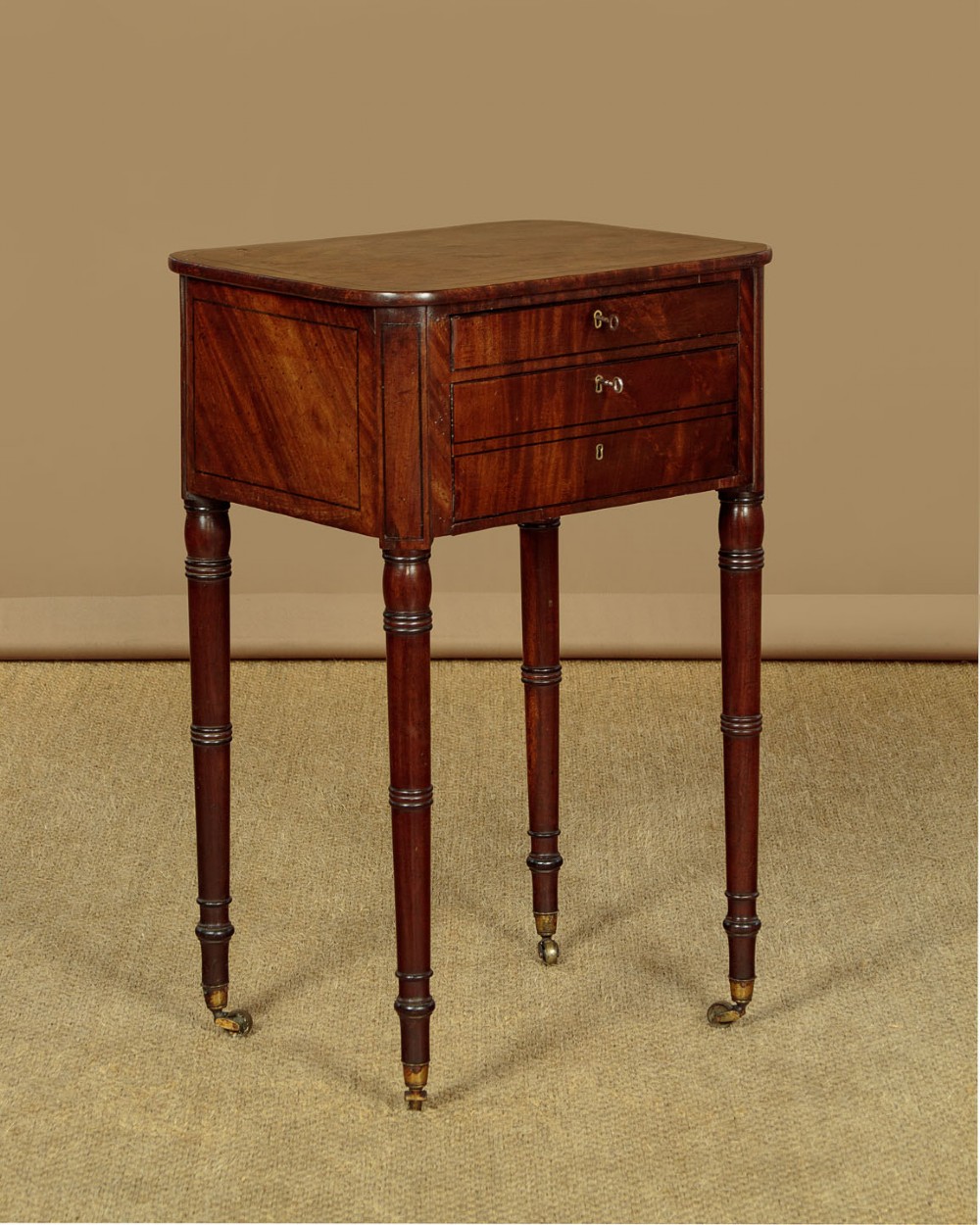 A George III mahogany side table featuring three shallow drawers, ring-turned legs on brass castors, and a rich wood grain finish.