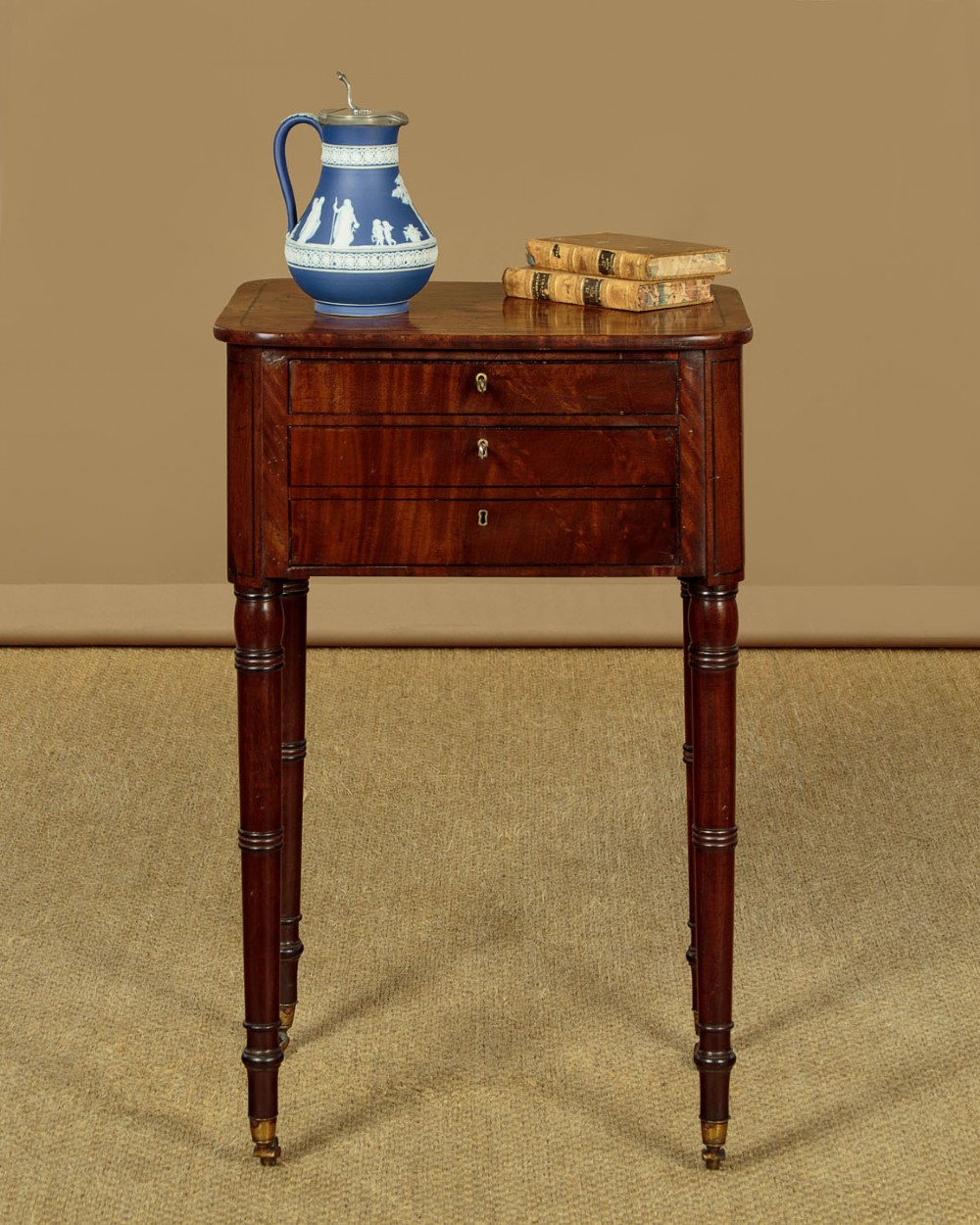 A George III mahogany side table featuring three shallow drawers, ring-turned legs on brass castors, and a rich wood grain finish. View II