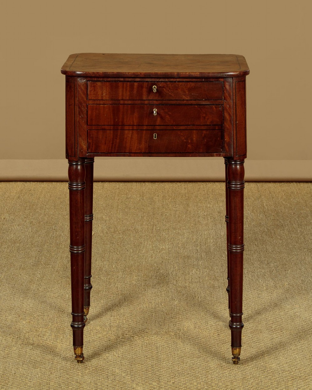 A George III mahogany side table featuring three shallow drawers, ring-turned legs on brass castors, and a rich wood grain finish. View III
