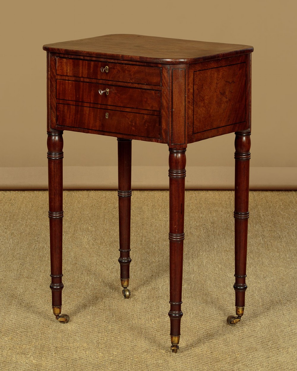 A George III mahogany side table featuring three shallow drawers, ring-turned legs on brass castors, and a rich wood grain finish. View IV
