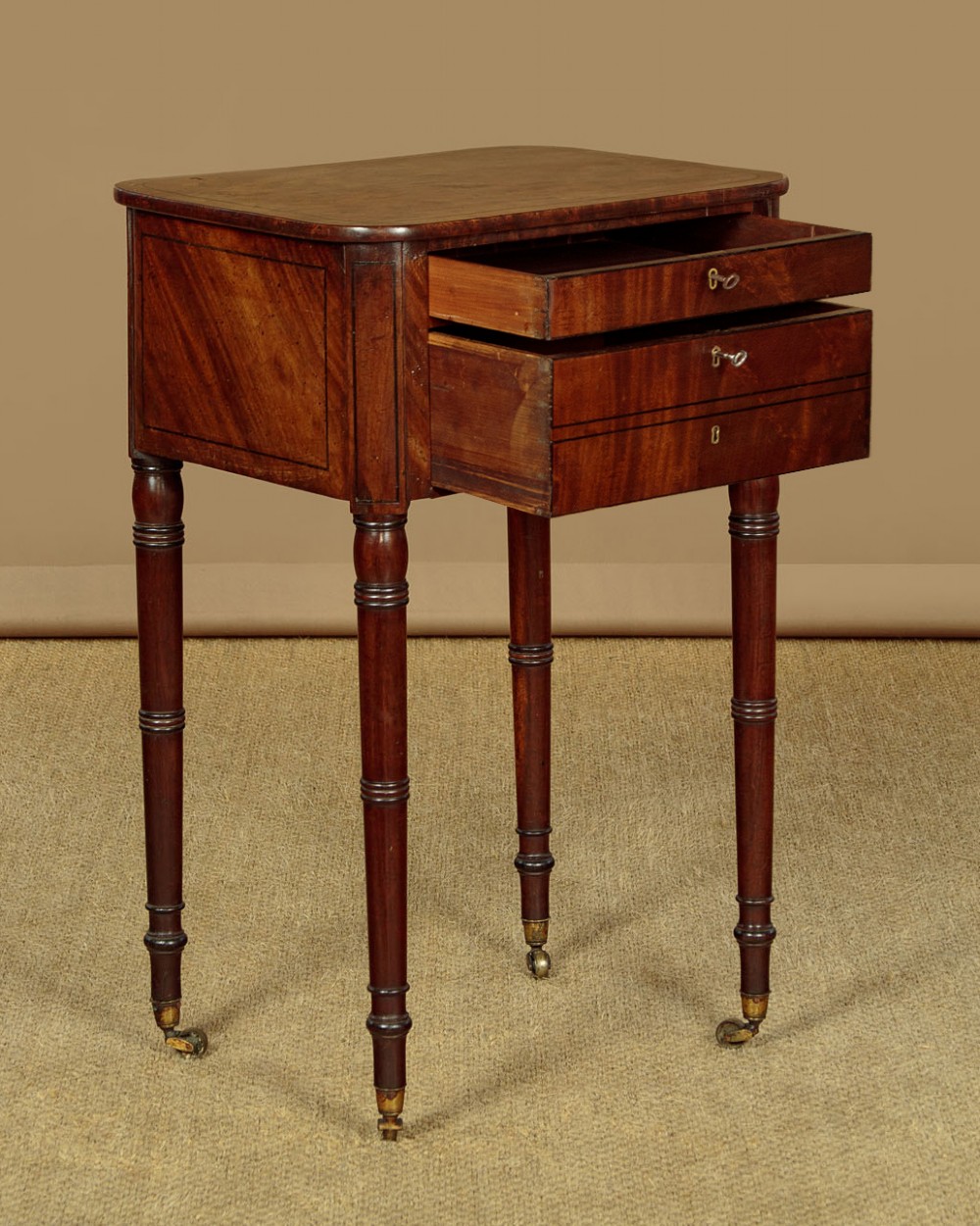 A George III mahogany side table featuring three shallow drawers, ring-turned legs on brass castors, and a rich wood grain finish. View V