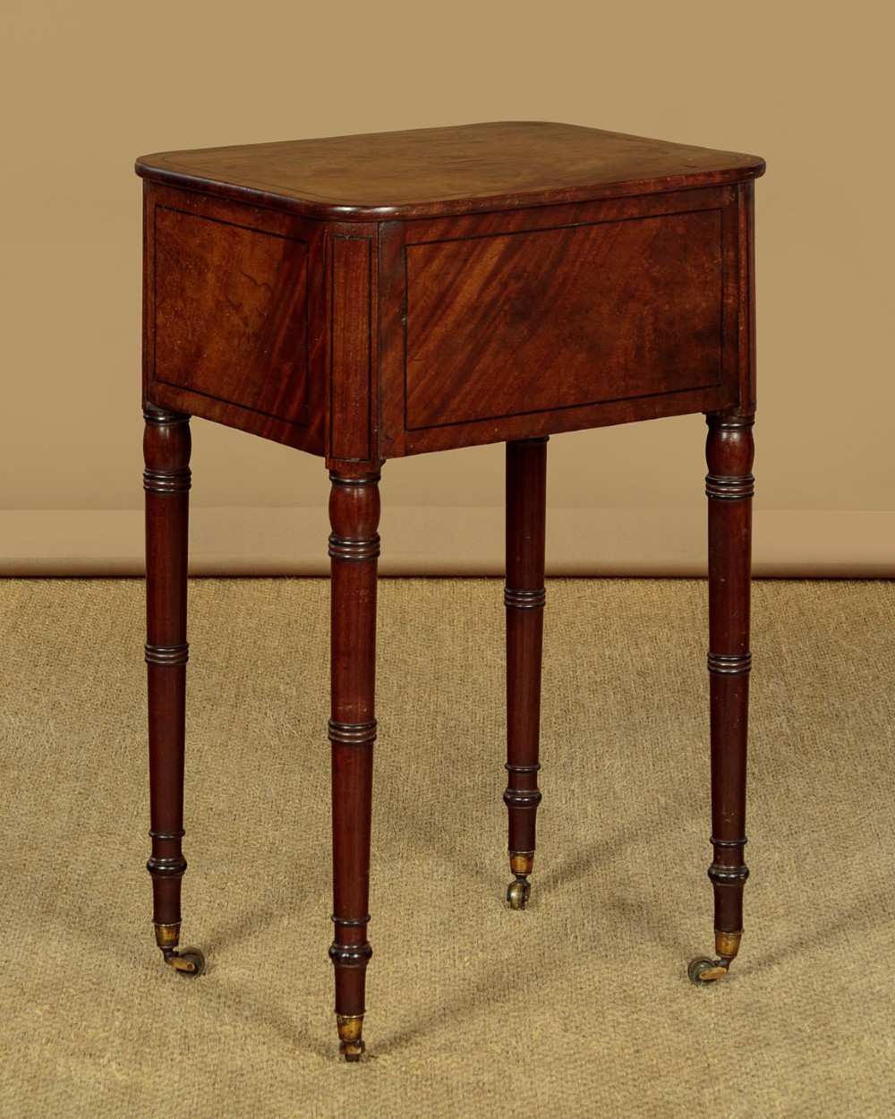 A George III mahogany side table featuring three shallow drawers, ring-turned legs on brass castors, and a rich wood grain finish. View VI
