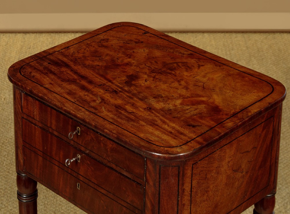 A George III mahogany side table featuring three shallow drawers, ring-turned legs on brass castors, and a rich wood grain finish. View VII