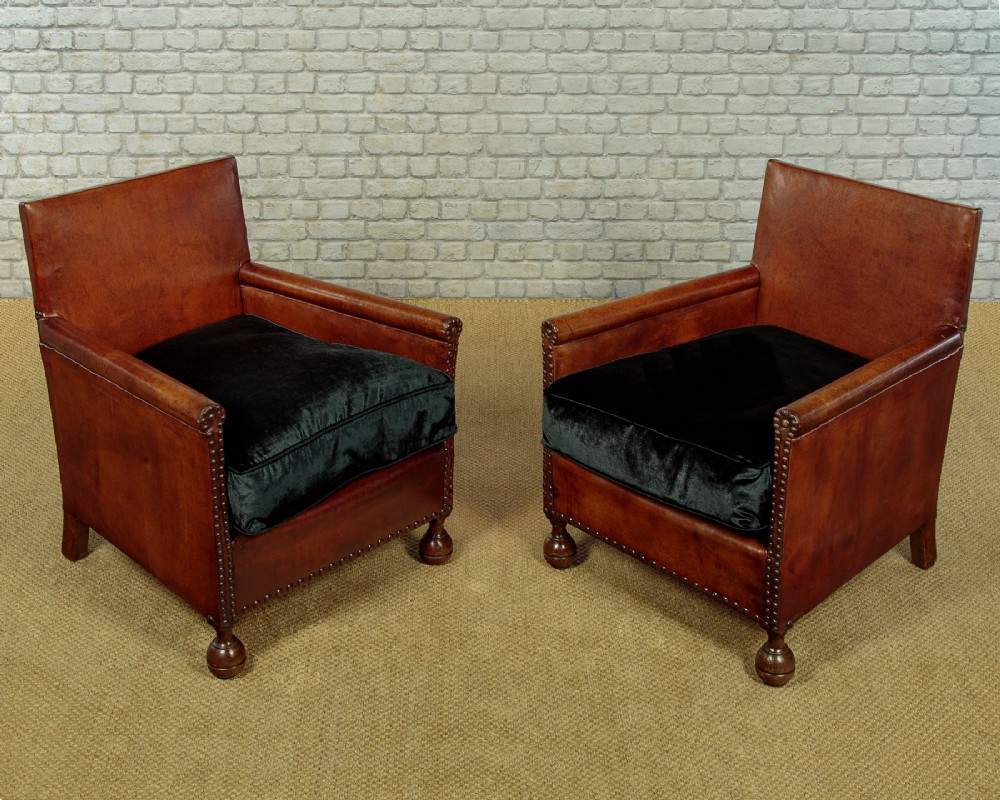 A pair of small 1920s brown leather armchairs featuring brass stud detailing, turned front legs, and dark velvet seat cushions.