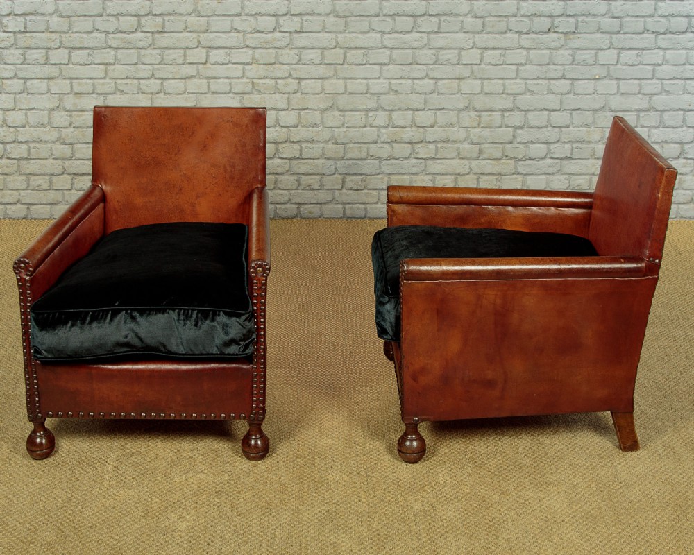 A pair of small 1920s brown leather armchairs featuring brass stud detailing, turned front legs, and dark velvet seat cushions. View II