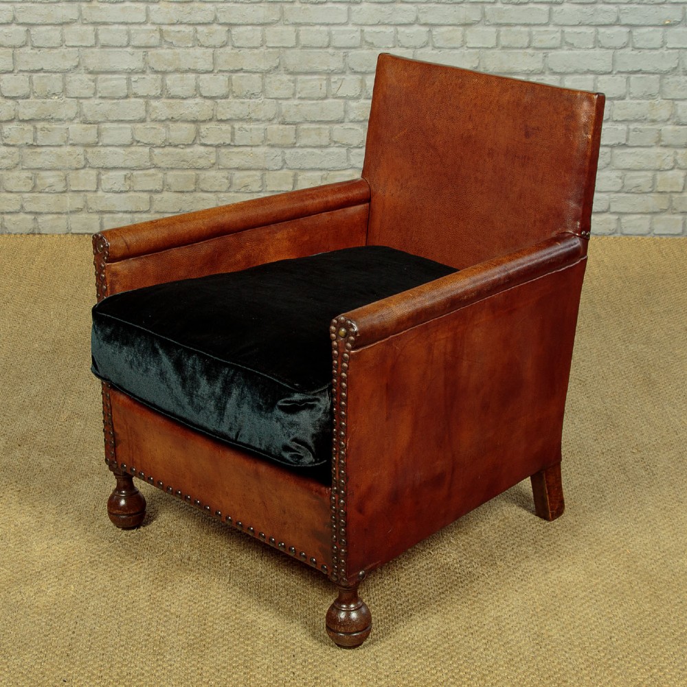 A pair of small 1920s brown leather armchairs featuring brass stud detailing, turned front legs, and dark velvet seat cushions. View V