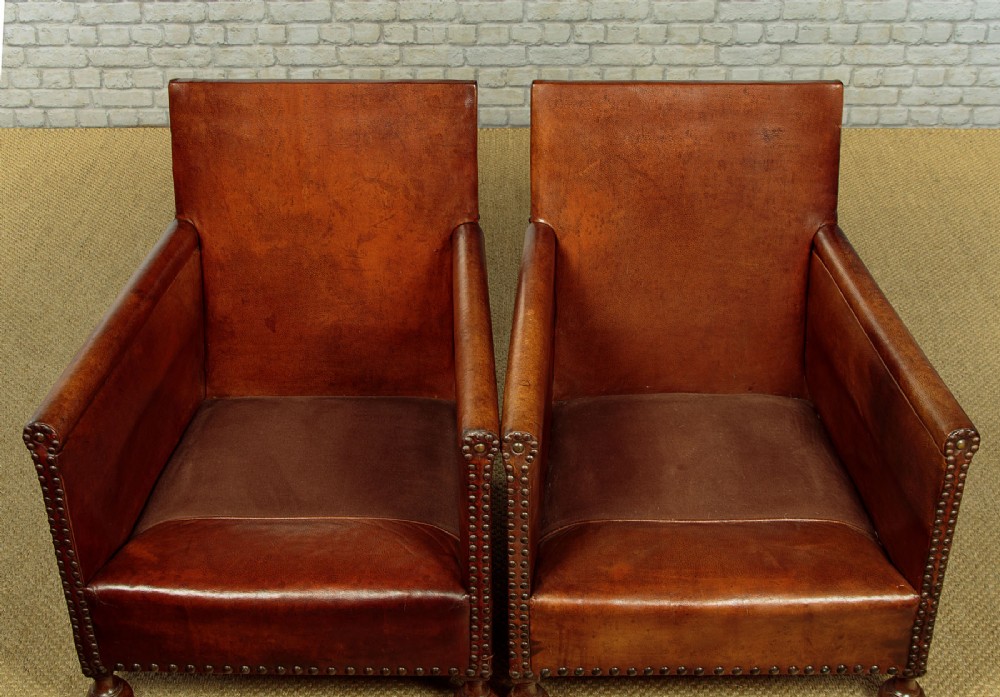 A pair of small 1920s brown leather armchairs featuring brass stud detailing, turned front legs, and dark velvet seat cushions. View VII
