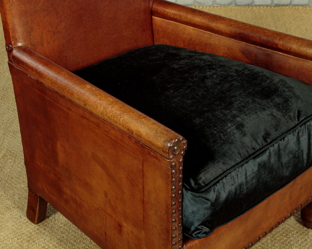 A pair of small 1920s brown leather armchairs featuring brass stud detailing, turned front legs, and dark velvet seat cushions. View X