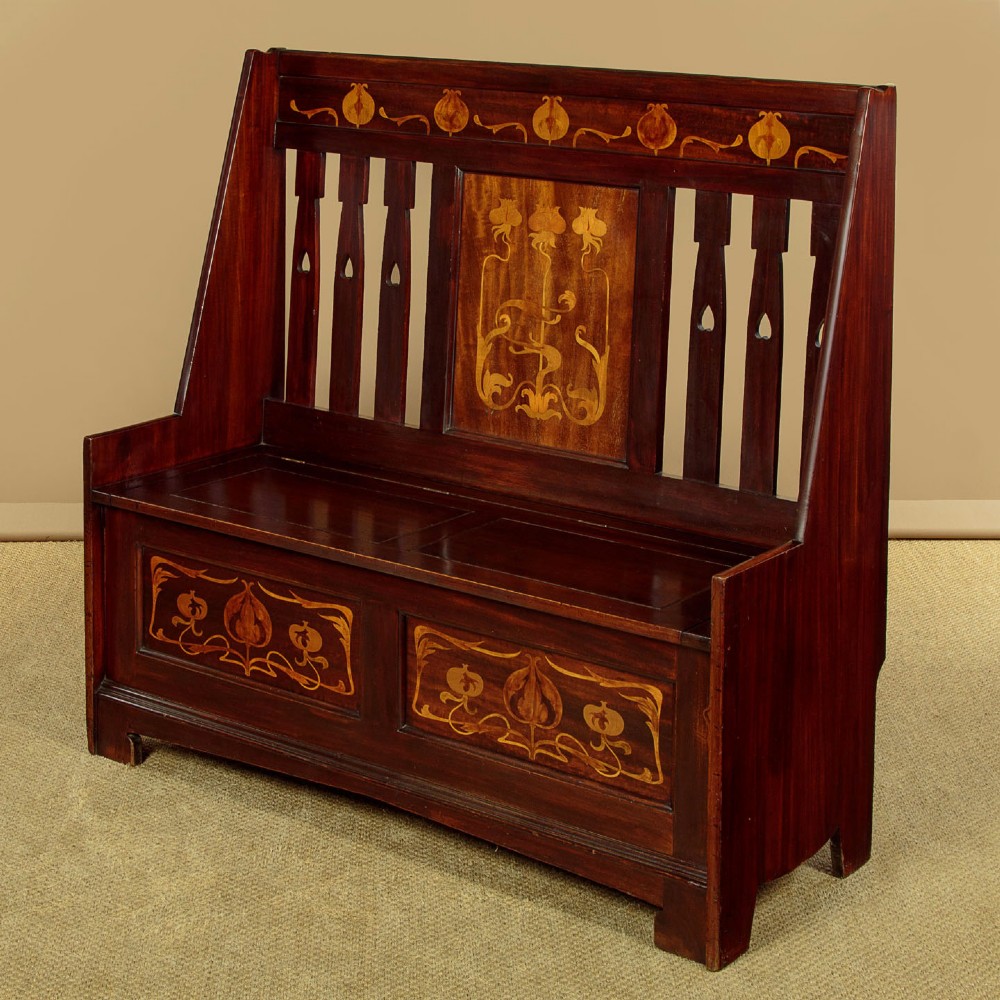 A dark mahogany high-back Arts and Crafts box settle featuring stylised floral marquetry inlay and vertical splat details above a hinged lift-up storage seat.