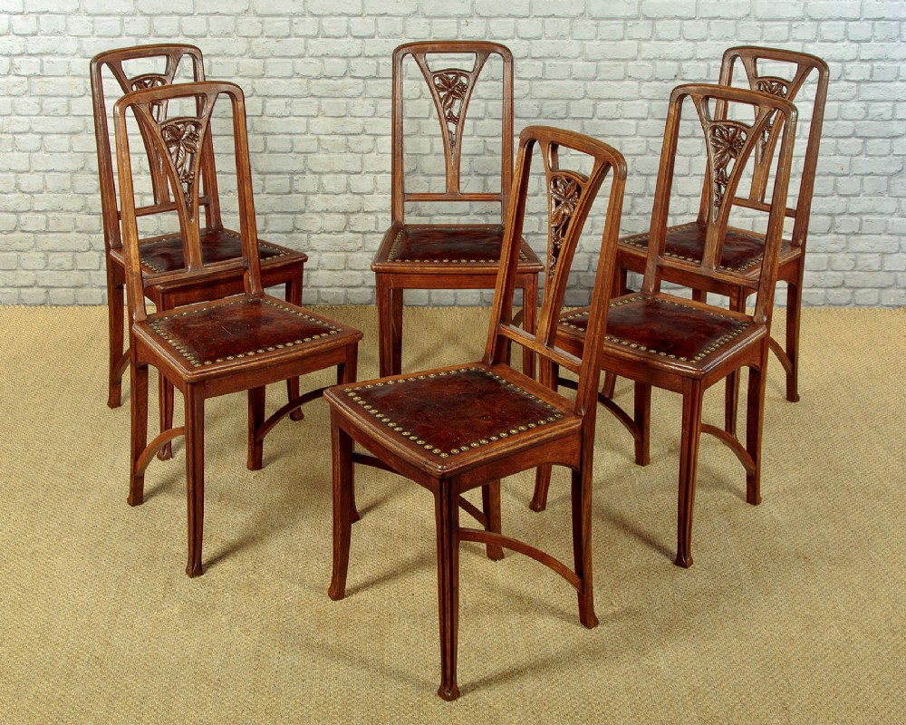 A set of six Art Nouveau oak dining chairs featuring carved botanical backrests and dark brown leather seats with brass stud trim.