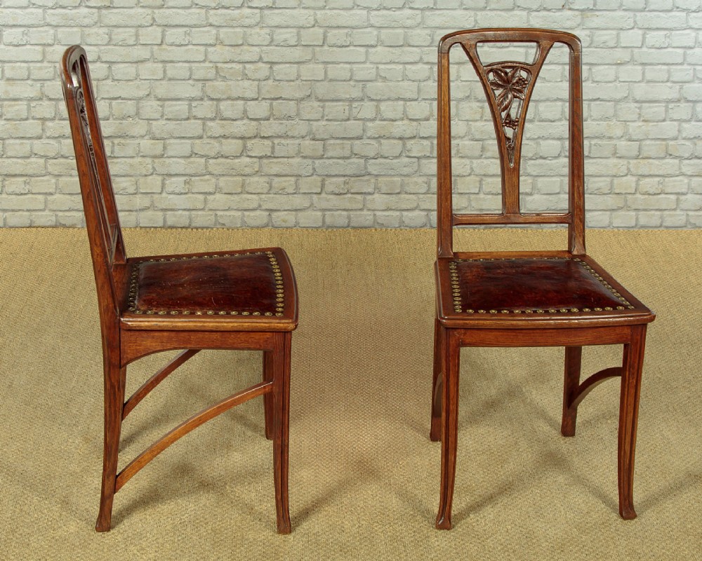 A set of six Art Nouveau oak dining chairs featuring carved botanical backrests and dark brown leather seats with brass stud trim. View II