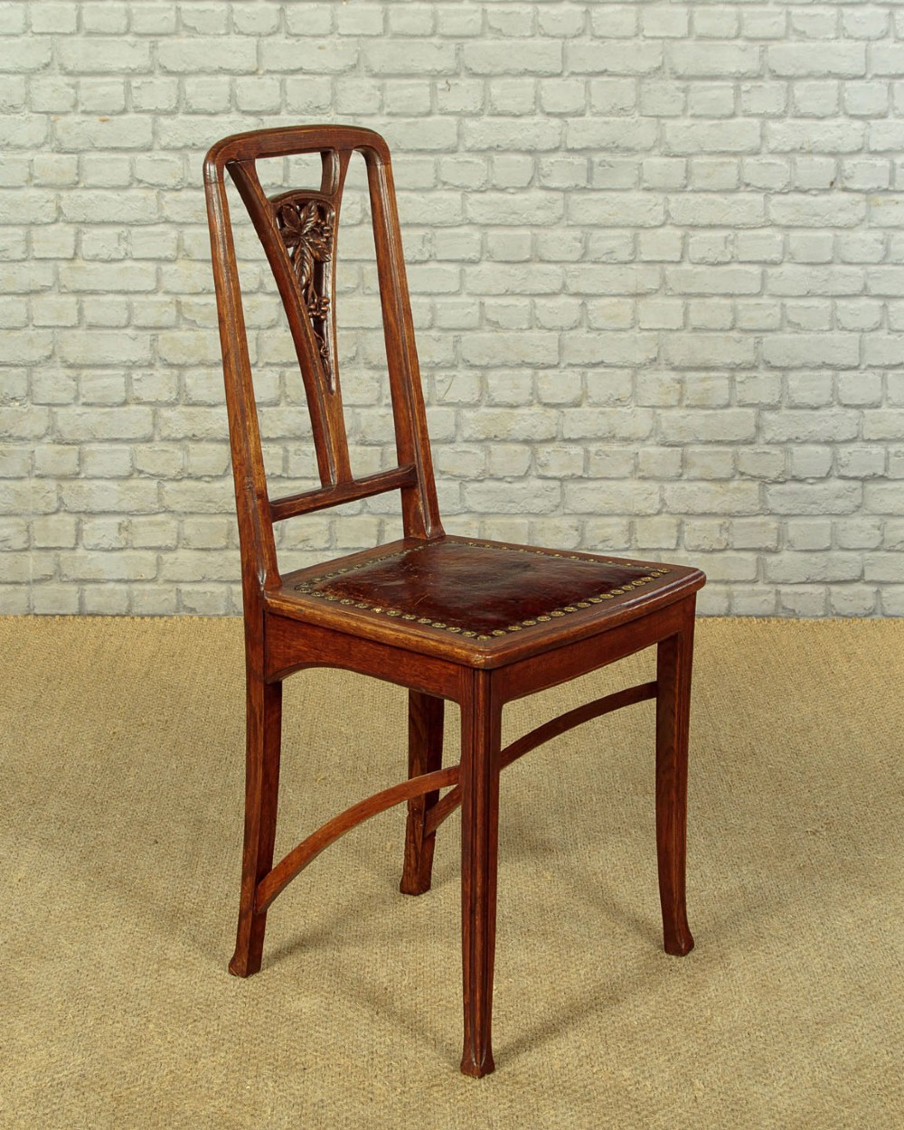 A set of six Art Nouveau oak dining chairs featuring carved botanical backrests and dark brown leather seats with brass stud trim. View V