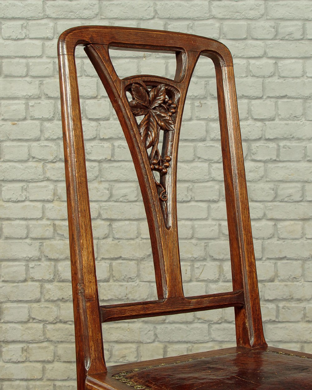A set of six Art Nouveau oak dining chairs featuring carved botanical backrests and dark brown leather seats with brass stud trim. View VI