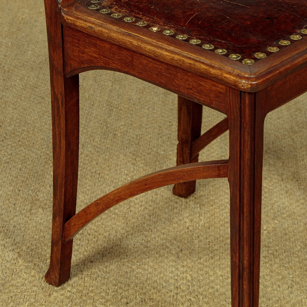 A set of six Art Nouveau oak dining chairs featuring carved botanical backrests and dark brown leather seats with brass stud trim. View XII