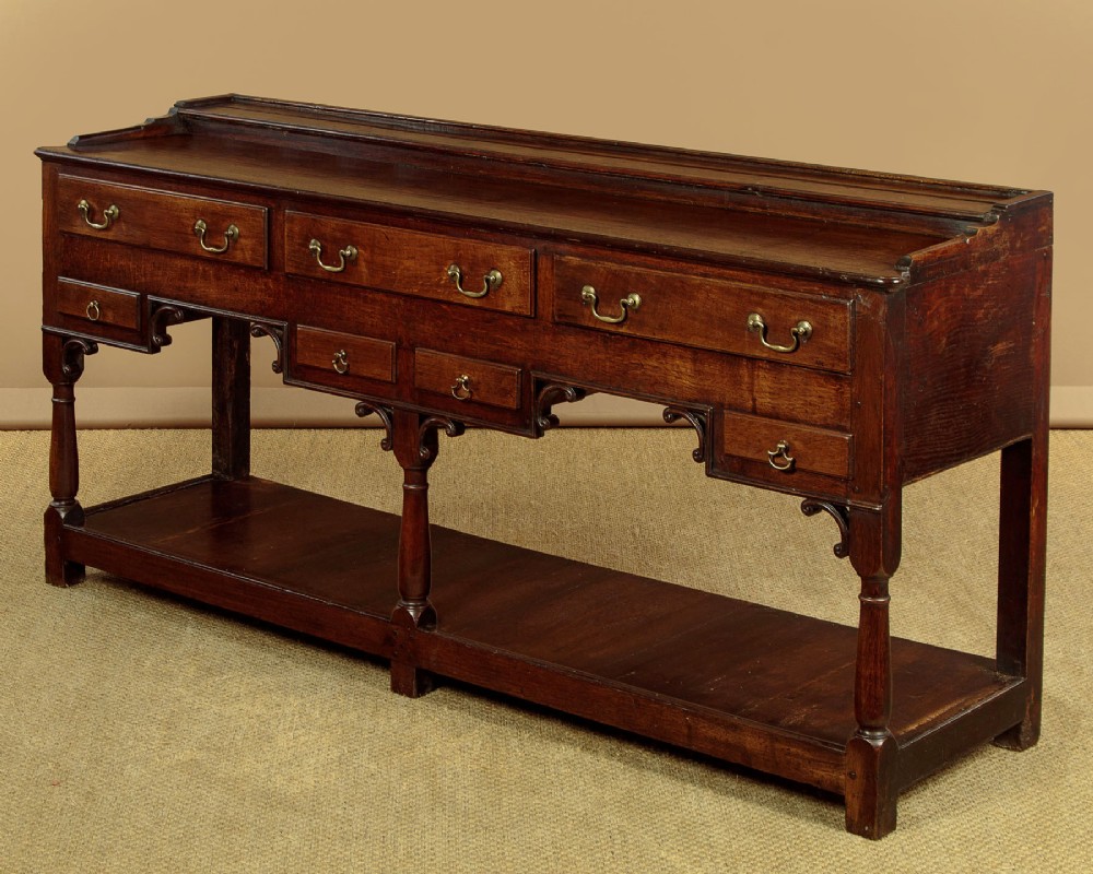 A George III dark oak potboard dresser base featuring three upper drawers, four frieze drawers, brass handles, shaped spandrels, and a low open shelf. View IV