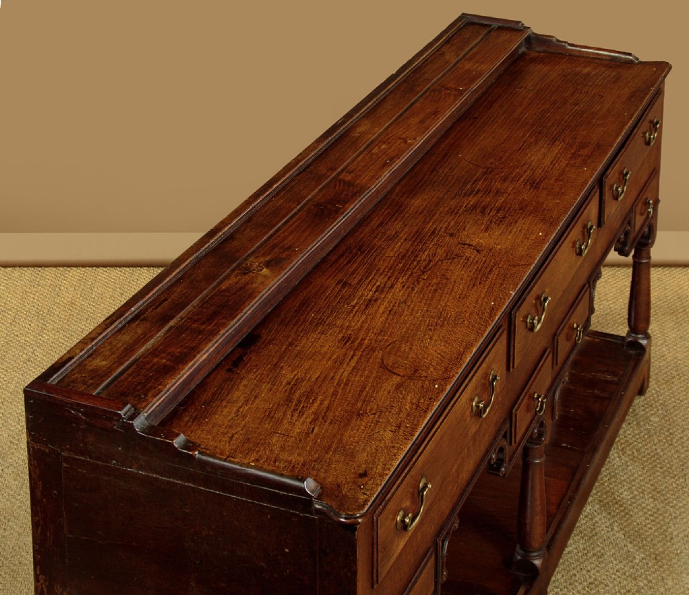 A George III dark oak potboard dresser base featuring three upper drawers, four frieze drawers, brass handles, shaped spandrels, and a low open shelf. View VI