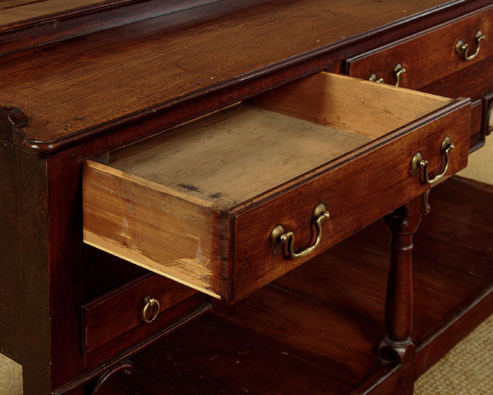A George III dark oak potboard dresser base featuring three upper drawers, four frieze drawers, brass handles, shaped spandrels, and a low open shelf. View IX