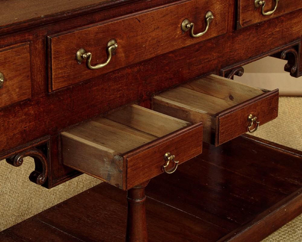 A George III dark oak potboard dresser base featuring three upper drawers, four frieze drawers, brass handles, shaped spandrels, and a low open shelf. View X