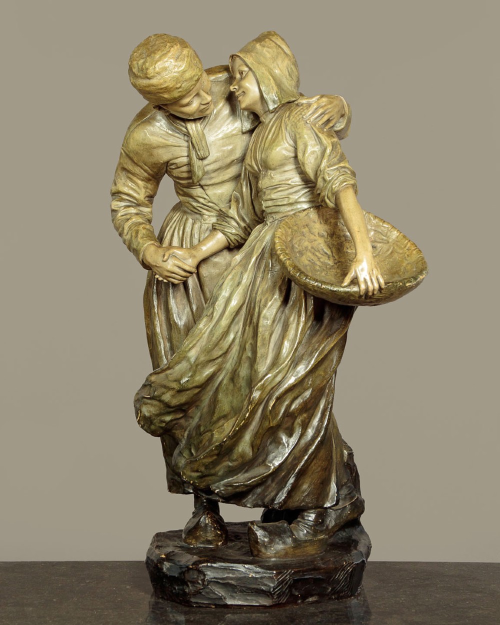 A patinated terracotta sculpture depicting a man and woman in traditional rural attire embracing while holding a woven basket.