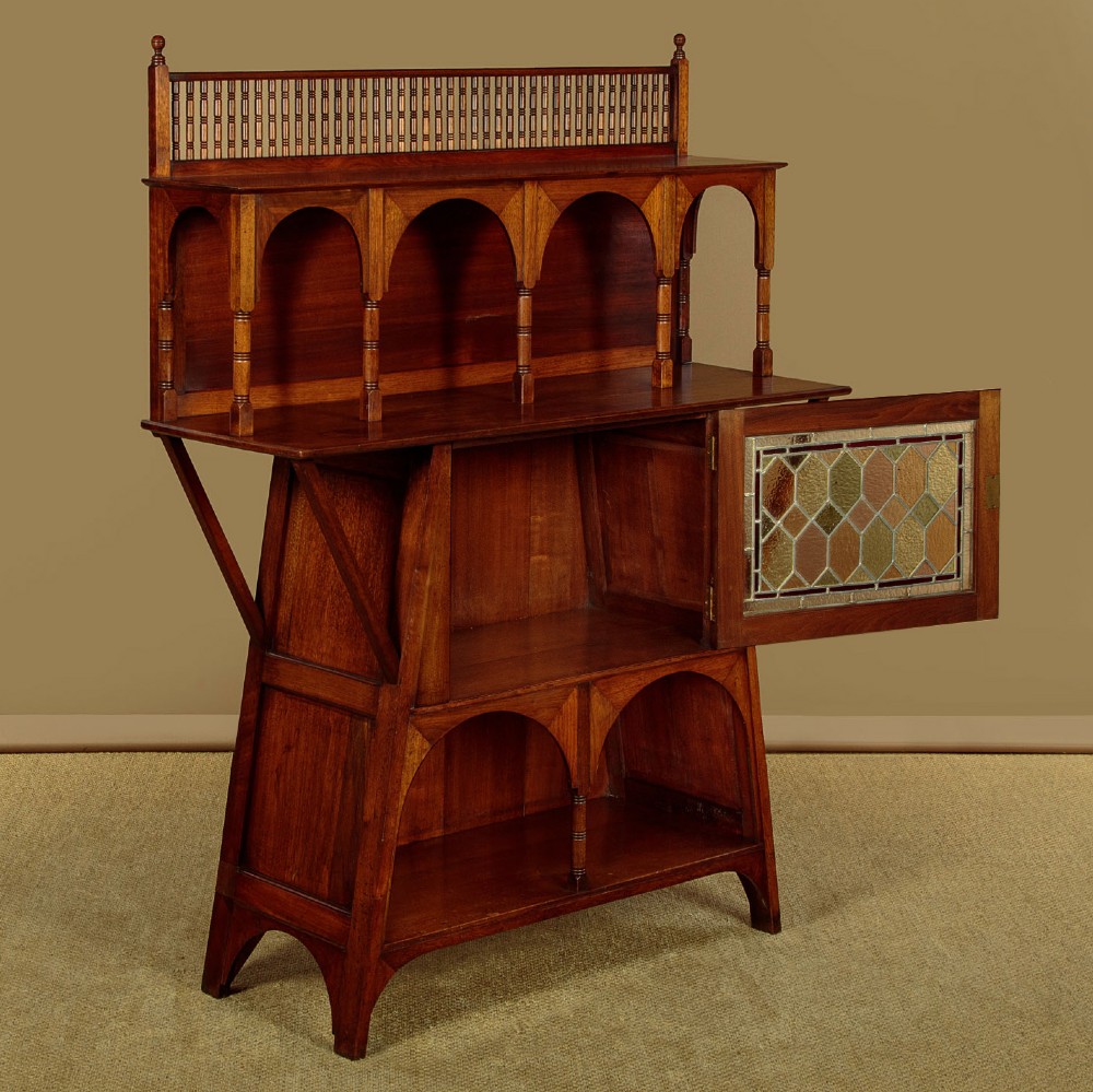 An Arts and Crafts oak side cabinet featuring an upper spindle gallery, arched open shelves, diagonal side struts, and a central cabinet door with leaded glass. View IV