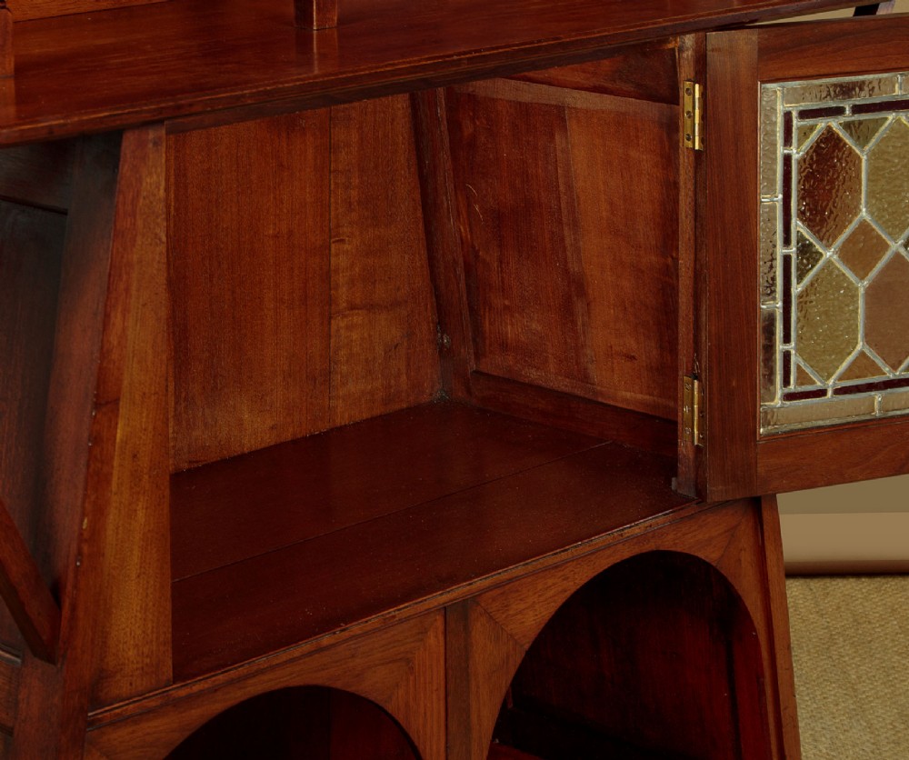 An Arts and Crafts oak side cabinet featuring an upper spindle gallery, arched open shelves, diagonal side struts, and a central cabinet door with leaded glass. View IX
