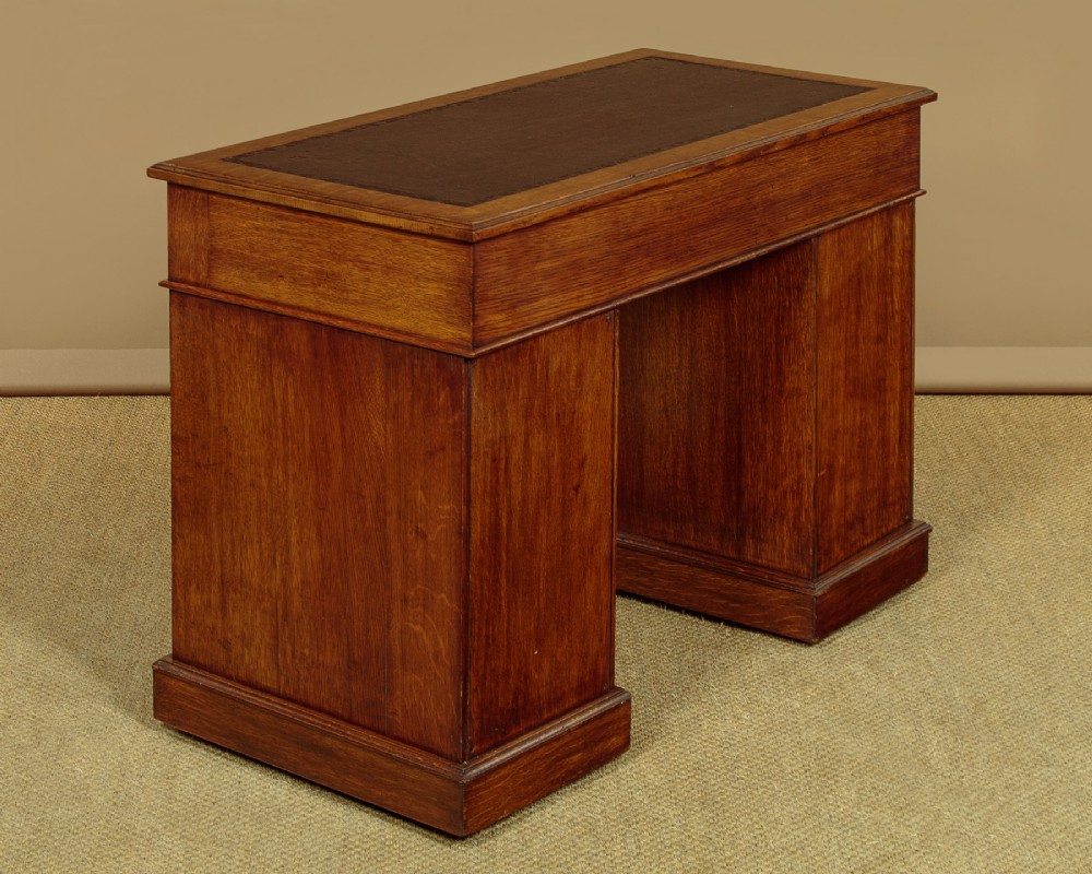 A late Victorian oak pedestal desk featuring a dark brown leather writing top, nine drawers with brass handles, and a solid plinth base. View X
