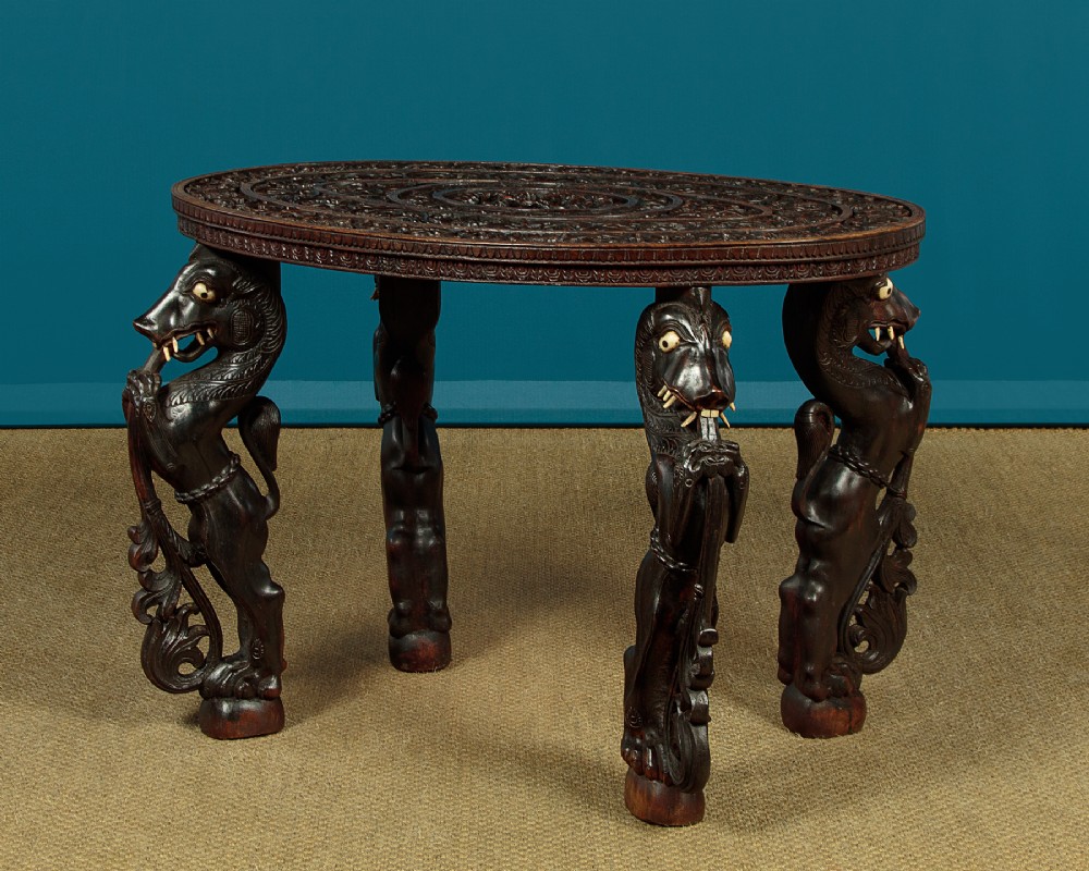 An oval dark hardwood Anglo-Indian coffee table featuring intricate top carvings and four legs sculpted as mythological beasts with inset white teeth.