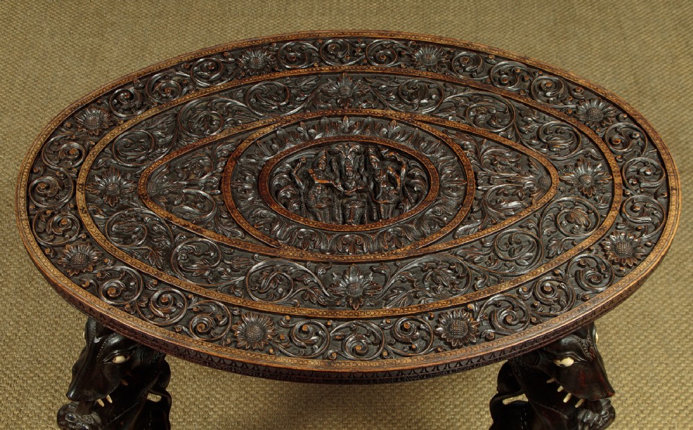 An oval dark hardwood Anglo-Indian coffee table featuring intricate top carvings and four legs sculpted as mythological beasts with inset white teeth. View II